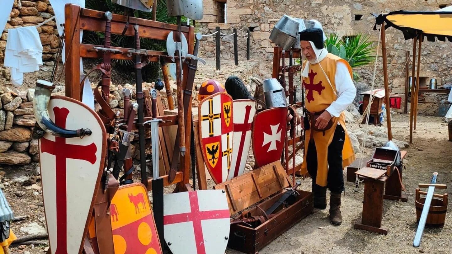 Recreador amb gonella groga davant una exposició d'escuts, espases i destrals al castell.
